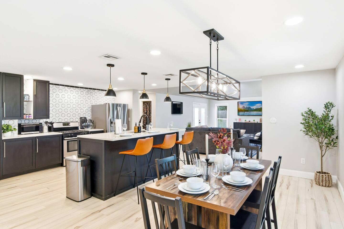 Dining Area – Wooden table set for six with modern lighting, black chairs, and open access to the kitchen with bar seating and stainless steel appliances.