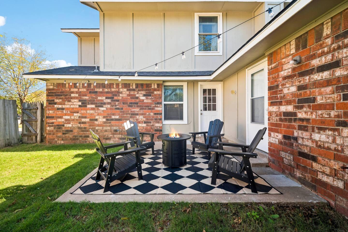 Backyard – Outdoor area with four black chairs around a fire pit on a black and white checkered patio, surrounded by string lights and grass.